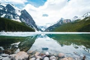 Mountain lake with clear water reflecting snow-capped peaks and pine forests under a cloudy sky.