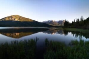 Calm lake with grassy shore reflecting forested hills and snow-capped mountains under clear sky