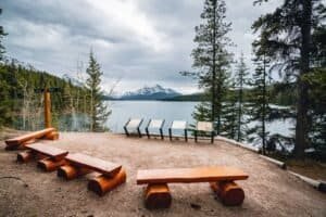 Lakeside viewpoint with wooden benches, informational boards, pine trees, lake, and snow-capped mountains.