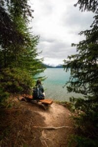 Person sitting on a wooden bench by a lake, surrounded by pine trees and mountains.