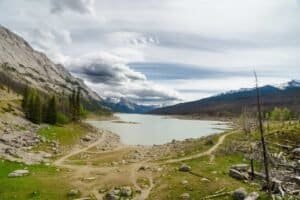Mountain lake with rocky shores, conifer trees, dirt paths, and snow-capped peaks under clouds.