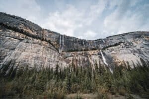 Rocky cliff with multiple narrow waterfalls above dense evergreen forest under cloudy sky