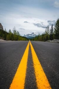 Paved road with double yellow lines leading to snow-capped mountain surrounded by trees under cloudy sky