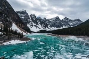 Partially frozen turquoise lake surrounded by snow-covered mountains and evergreen forests.