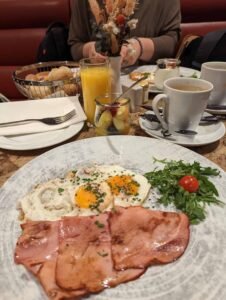 Breakfast plate with fried eggs, ham, greens, cherry tomato, orange juice, fruit salad, and coffee.