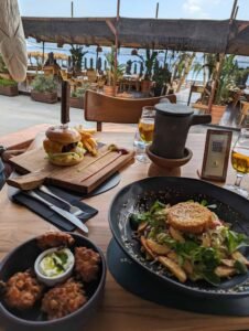 Outdoor wooden table with cheeseburger, fries, salad with patty, fried food, beer, and teapot.
