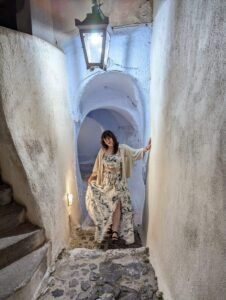 Woman in floral outfit stands in narrow cobblestone alley with white textured walls and arched ceiling.