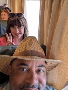 Man wearing a woven straw hat in foreground, woman behind making a peace sign indoors.