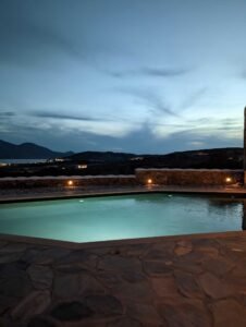 Outdoor illuminated swimming pool at dusk with stone patio and distant hills under cloudy sky.