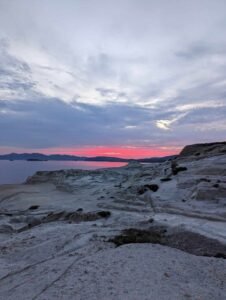 Rocky coastal landscape at dusk with calm sea, distant hills, and pink to grayish-blue sky gradient.