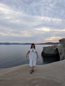 Woman in white dress and black sandals stands on rocky shore with calm water and cloudy sky.