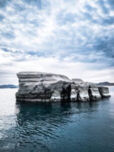 Large light-colored layered rock formation in calm water under a cloudy sky with distant hills.