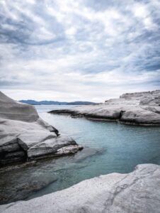 Calm coastal water surrounded by smooth pale rock formations and distant hills under a cloudy sky.