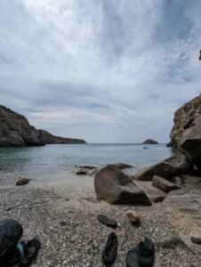 Rocky beach with pebbles, black water shoes, calm sea, and cliffs under a cloudy sky.