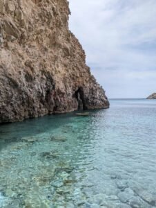 Rocky cliff with a cave extends into clear turquoise water under a partly cloudy sky.