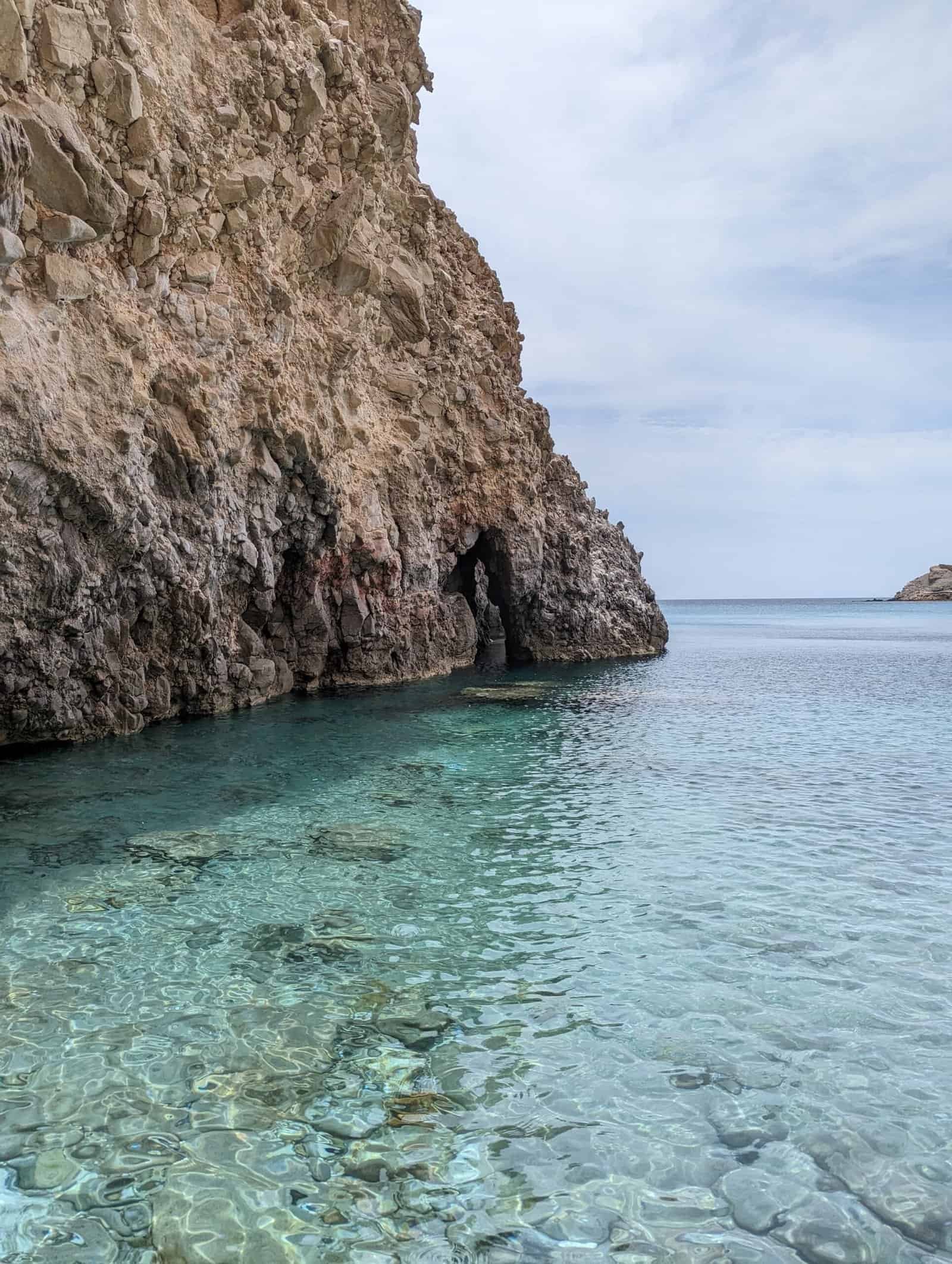 Rocky cliff with turquoise water and sea cave Rocky cliff with a cave extends into clear turquoise water under a partly cloudy sky.