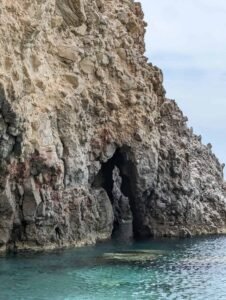 Rocky coastal cliff with natural arch over clear blue-green water under cloudy sky.