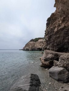Rocky coastline with clear calm water and pebble beach under an overcast sky.