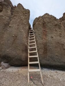 Wooden ladder leans against narrow gap between two tall rock formations under overcast sky.