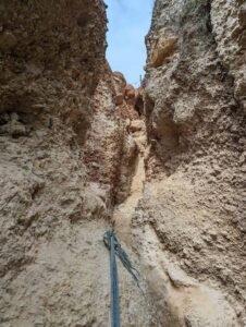 Narrow dirt path through steep rocky cliffs with a blue rope anchored for climbing.