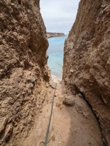Narrow rocky path between cliffs leads down to calm blue sea with rope on the ground.