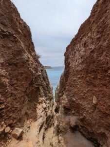 Narrow rocky crevice opening to calm blue sea under overcast sky with distant land visible.