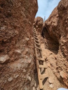 Wooden ladder leaning against reddish rock walls in a narrow outdoor crevice with a dirt floor.