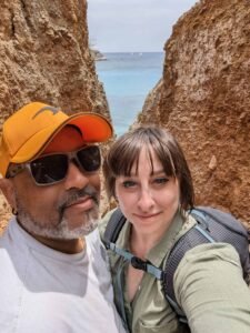Two people taking a selfie between cliffs with ocean and sailboats in the background.