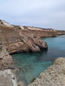 Rocky beach with clear turquoise water, surrounded by cliffs, with people on shore and swimming.