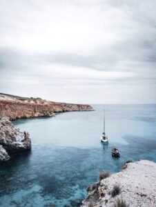 Coastal scene with clear blue water, rocky shore, a sailboat, a dinghy, and cloudy sky.