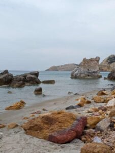 Rocky sandy beach with large boulders in calm shallow water and hilly coastline under overcast sky