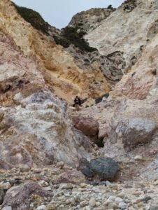 Person standing on rocky slope surrounded by multicolored cliffs with sparse vegetation under overcast sky