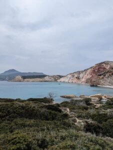 Coastal landscape showing calm blue sea, rugged cliffs, green shrubbery, and cloudy sky.