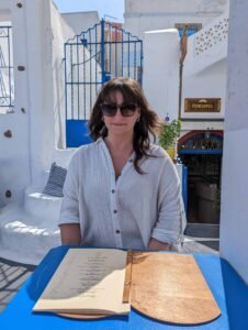 Woman in sunglasses and white blouse sitting at blue table with open menu outdoors.