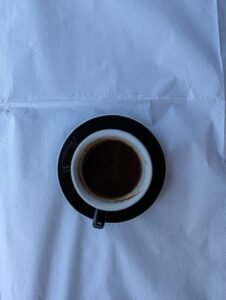 White cup with black handle and saucer containing dark liquid on textured white cloth.