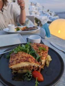 Two slices of baked layered dish with arugula and cherry tomato on a black plate at a table.