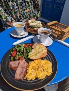 Breakfast on a blue table with scrambled eggs, ham, salad, toast, coffee, and feta salad.