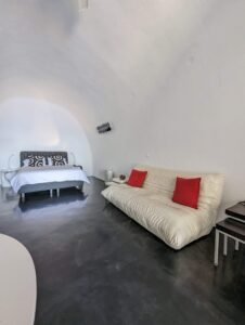 Minimalist bedroom with curved white ceiling, dark floor, gray bed, white sofa with red pillows, and side tables.