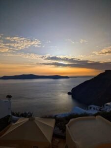 Sunset over calm sea with silhouetted cliffs, beige umbrellas, and white buildings in foreground.