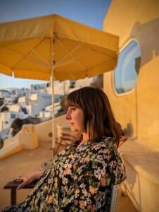 Woman in floral shirt sitting under a yellow umbrella on a terrace with white buildings in background.