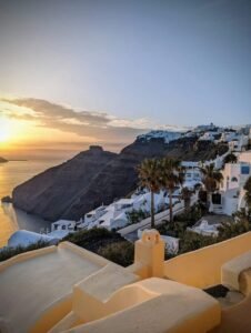 Sunset over a coastal cliffside village with white cubic buildings and palm trees.