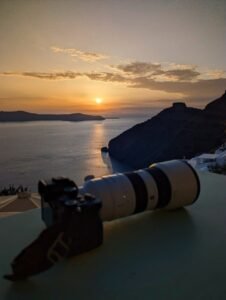 Camera with telephoto lens resting on a surface overlooking a coastal sunset scene.
