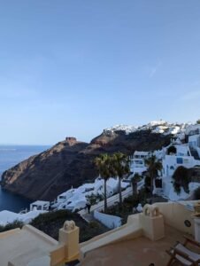 Coastal hillside town with white buildings, palm trees, rocky cliffs, and sea under clear sky