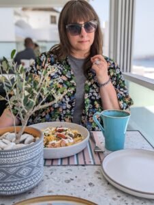 Person in floral shirt and sunglasses at table with food, blue mug, empty plate, and potted plant.