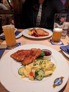 Two plates with breaded schnitzel, pasta salad, red sauce, two glasses of beer, and a salt grinder.
