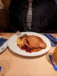 Plate with breaded schnitzel, spaetzle with crispy onions, brown gravy, cranberry sauce, and orange slice on a wooden table.