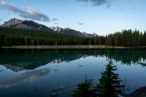 Calm lake reflecting evergreen trees and snow-capped mountains under a partly cloudy sky