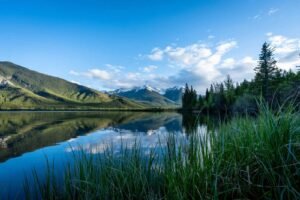 Calm lake reflecting green hills, pine trees, snow-capped mountains, and blue sky with clouds.