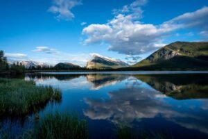 Lake with mountain reflections, cloudy blue sky, and grasses along shoreline
