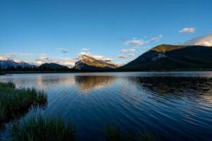 Lake reflecting mountains under a partly cloudy sky with grasses at the water's edge at sunset
